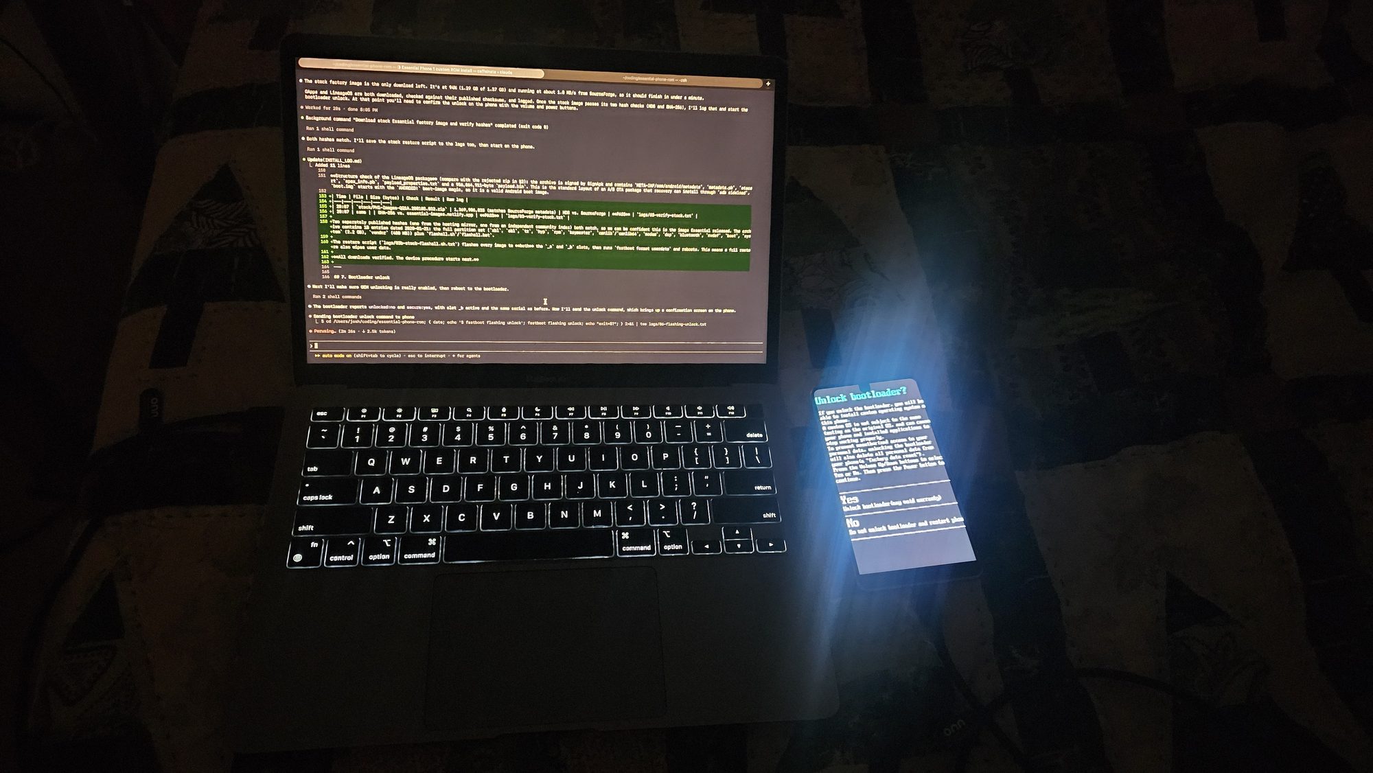 A MacBook Air in a dark room showing Claude Code's terminal output, next to an Essential Phone displaying the "Unlock bootloader?" confirmation screen